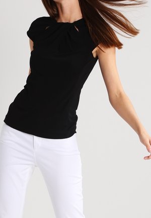 Vrouw draagt een mouwloze zwarte top met geplooide halslijn en witte broek, haar dat naar de zijkant stroomt tegen een effen lichte achtergrond.