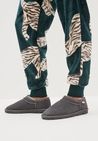 Pantofole di camoscio grigio con profilo basso, dettagli cuciti e suole di gomma strutturate, abbinate a pantaloni del pigiama verdi con motivi di tigre.
