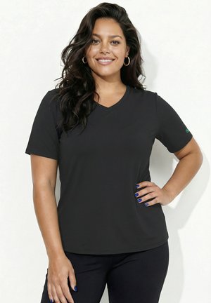 Lächelnde Frau mit langen dunklen Haaren, die ein schwarzes T-Shirt mit V-Ausschnitt und schwarze Hose trägt, eine Hand an der Hüfte, blaue Nagellackierung, goldene Creolen.