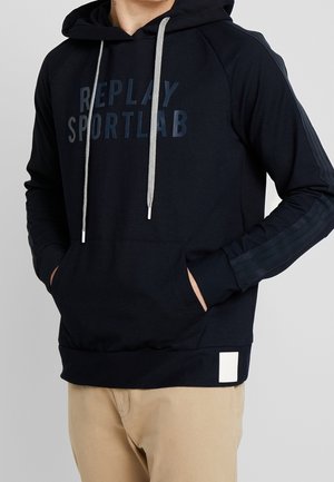 Mand iført en sort hættetrøje med grå snore, "REPLAY SPORTLAB" tekst på brystet, hænder i forlommerne, beige bukser.