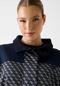 Marineblauwe trui met een hoge kraag, met een patroon aan de voorkant in wit en marineblauw, afgewerkt met een logo en metalen accenten.