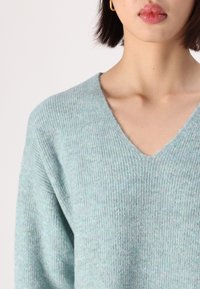 Pull en fine maille bleu clair avec un décolleté en V. Il présente une texture douce et une coupe ample, mettant en valeur son style confortable et décontracté.