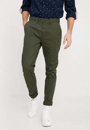 Mand iført op-rullede olivengrønne chinos med en navyblå prikket skjorte og hvide sneakers, går fremad.
