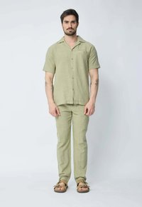 Chemise en lin vert clair à manches courtes avec un bouton sur le devant, associée à un pantalon en lin assorti, agrémentée de sandales marron. Texture lisse.