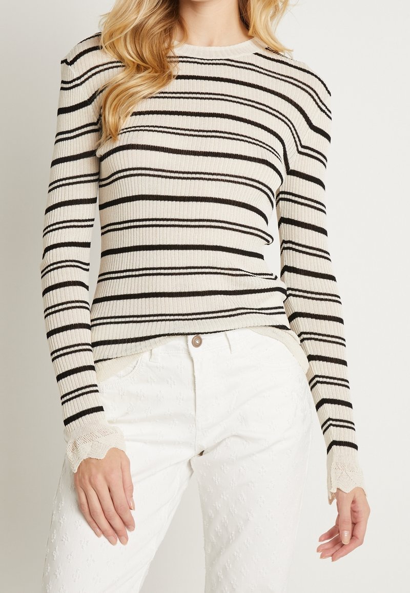 Suéter de rayas negro y crema con canalé y mangas largas, que presenta detalles de encaje en los puños, llevado sobre pantalones blancos texturizados.