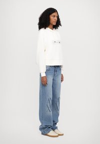 Hoodie bianco cropped con testo stampato, abbinato a jeans blu larghi con dettagli decorativi. Scarpe chiare completano l'outfit.