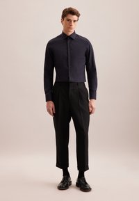 Camicia blu navy con design accorciato, abbinata a pantaloni neri su misura con risvolti e scarpe nere. Texture liscia in tutto.