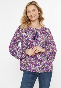 Lila blumiges Blusenshirt mit luftigen langen Ärmeln, einem gerafften Halsausschnitt mit einem Bindeband und mehrfarbigen Blumenmustern in Rosa- und Weißtönen.