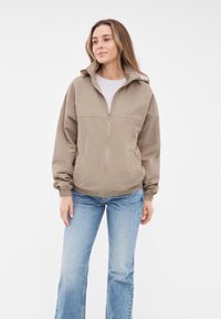 Giacca beige con zip, colletto alto, polsini elasticizzati e vestibilità rilassata, abbinata a jeans a campana blu chiaro. Design semplice senza motivi.