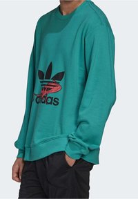 Teal-sweatshirt, jossa on musta ja punainen grafiikka sekä Adidas-logo ja kolme raitaa -design. Valmistettu pehmeästä kankaasta, jossa on ribbireunat hihoissa ja helmassa.