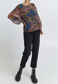 Lettet, gennemsigtig bluse med et livligt blomsterprint i pink, grøn og blå; rund halsudskæring; parret med sorte slanke bukser og ankelstøvler.