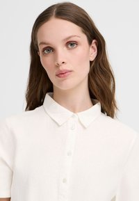 Camicia bianca a maniche corte con colletto e bottoni sul davanti, realizzata in tessuto testurizzato, caratterizzata da un design classico e dettagli di cucitura discreti.