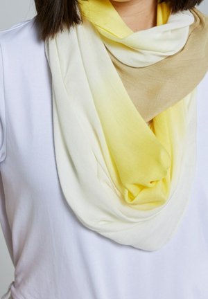 Femme portant un foulard doux et léger aux nuances de blanc, jaune et beige sur un haut blanc uni, mettant en valeur la texture et la chute du foulard.