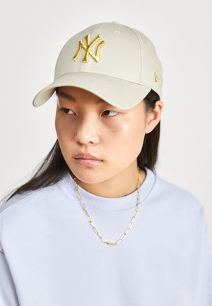 Ung kvinde iført beige baseballcap med gyldent New York Yankees-logo og lyseblå skjorte, der kigger til venstre.
