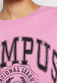 Sudadera rosa con texto impreso en negro y un diseño de logo circular. La tela parece suave, con cuello redondo y corte estándar.
