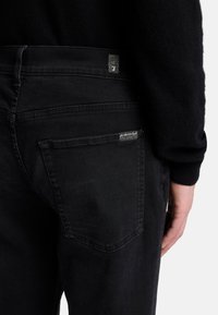 Jeans in denim nero con una texture liscia, taglio dritto e un design distintivo delle tasche posteriori con etichette. Cuciture visibili e dettagli di ferramenta.