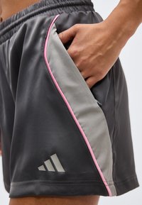 Χέρι στην τσέπη μαύρου και γκρι αθλητικού σορτς με ροζ περιθώριο και λογότυπο Adidas στο αριστερό πόδι.
