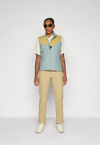 Veston léger sans manches en bleu et jaune, avec zip à l'avant, associé à une chemise légère et un pantalon beige. Les chaussures sont de couleur crème.