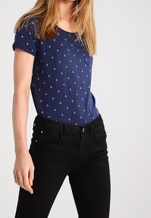 Kobieta w granatowym t-shircie z małymi białymi wzorami kotwic oraz czarnych dżinsach z wysokim stanem na jednolitym tle.