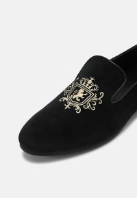 Chaussure en velours noir à enfiler avec un blason brodé en or représentant un lion couronné et des éléments décoratifs ornés sur la partie avant supérieure.
