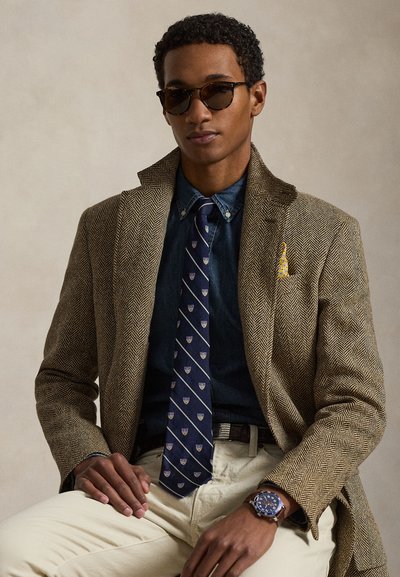 Uomo con occhiali da sole, blazer marrone a spina di pesce, cravatta blu navy a fantasia, camicia di jeans, pantaloni beige e orologio da polso seduto contro uno sfondo neutro.