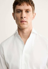 Chemise blanche à col écarté, avec une texture lisse et sans motifs visibles, portée par une personne aux cheveux courts.