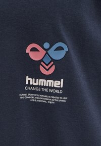 Un tissu bleu marine avec une surface texturée présente un logo central en rose et bleu. Le texte en dessous indique "HUMMEL CHANGE THE WORLD."