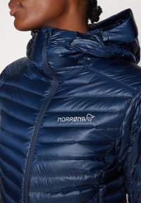 Norrøna puf jakna u tamnoplavoj boji, s kapuljačom, kvadratnim dizajnom i sjajnom teksturom. Logo prikazan na prsima.