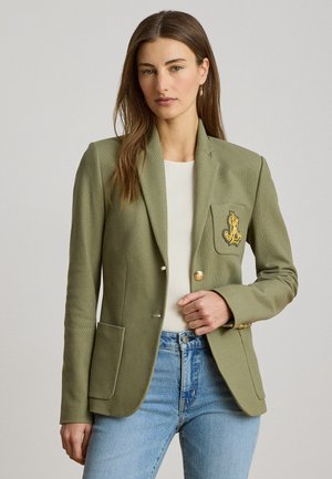 Vrouw draagt een olijfgroene blazer met gouden knopen en borduursel op de zak, een witte geribde top en lichtblauwe spijkerbroek.