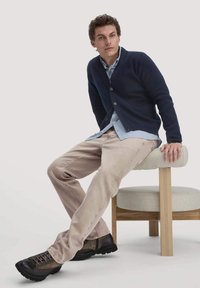 Marineblauwe knopenop cardigan over een lichtblauw shirt, gecombineerd met beige broek en donkerbruine schoenen. Zittend op een licht houten stoel.