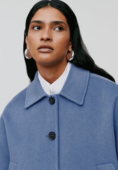 Cappotto di lana azzurro chiaro con colletto a punta, cinque bottoni neri e taschino sul petto, abbinato a una camicia bianca sotto.