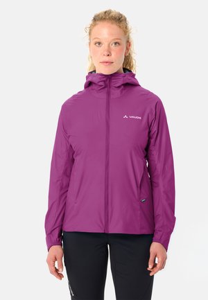 Frau mit lockigem Haar, die eine lila Jacke mit Reißverschluss und Kapuze sowie schwarze Hosen trägt, steht vor einem schlichten weißen Hintergrund.