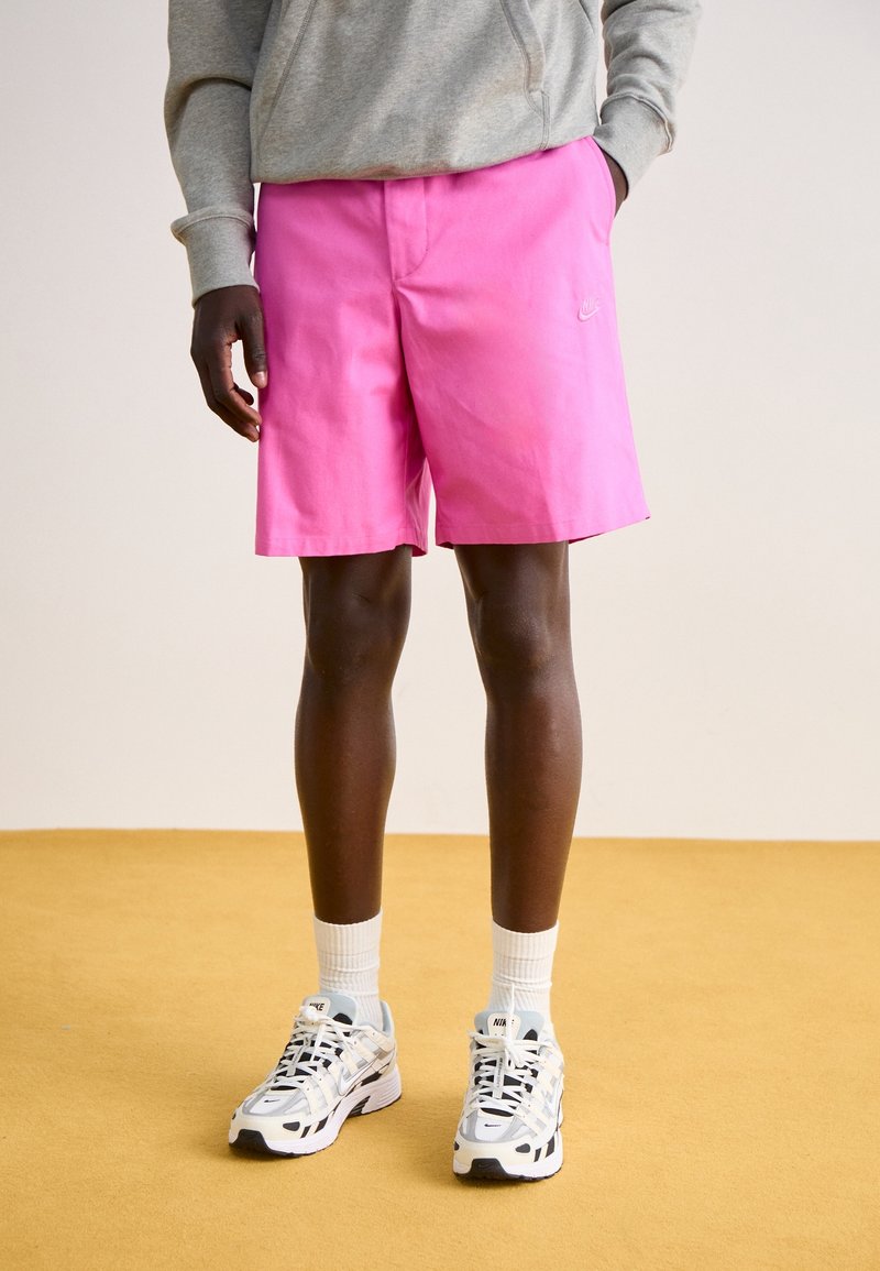 Rosa shorts med en platt frontdesign och en liten logo, kombinerat med vita sneakers och ankelsockor, på en gul golvyta.