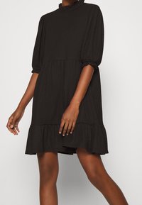 Abito nero realizzato in tessuto strutturato. Presenta un alto colletto arricciato, maniche a sbuffo e un orlo a balze. Design loose fit, lunghezza fino al ginocchio.