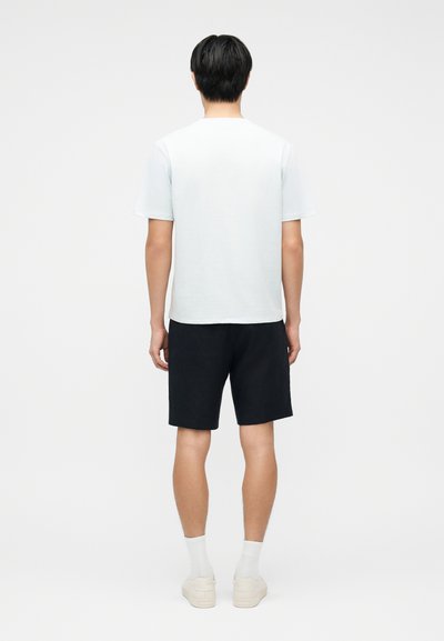 Jeune homme debout, de dos, portant un t-shirt blanc uni, un short noir, des chaussettes blanches et des baskets blanches sur un fond blanc.