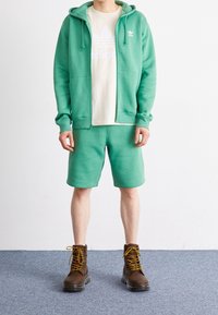 Uomo con felpa verde e pantaloni corti abbinati sopra una maglietta Adidas bianca, indossa stivali marroni con lacci e si trova su un tappeto grigio.