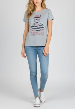 Camiseta gris con un estampado gráfico de un personaje que lleva gafas y un sombrero. Combinada con vaqueros ajustados azul claro y zapatillas blancas.