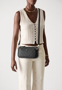 Mustra mustrist crossbody kott, kuldsete detailide ja logoga. Kantakse koos beeži kudumise vestiga ja heledate pükstega.