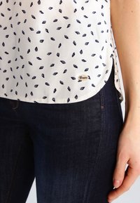 Camicia bianca con piccoli motivi a foglia navy, orlo arrotondato e un'etichetta dorata del marchio. Abbinata a jeans in denim blu scuro.