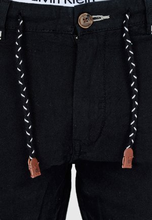 Black trousers with a button closure, featuring a drawstring with brown leather accents and a textured fabric. Stitching detail visible.