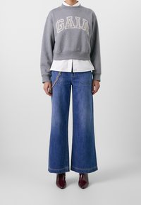 Grijze sweatshirt met borduursel "GAIA", witte blouse eronder, brede blauwe spijkerbroek en kastanjebruine puntige laarzen.