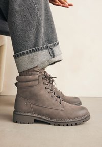Botas de tobillo marrones hechas de material sintético texturizado, con cordones, ojetes de metal y una suela resistente con un dibujo pronunciado.