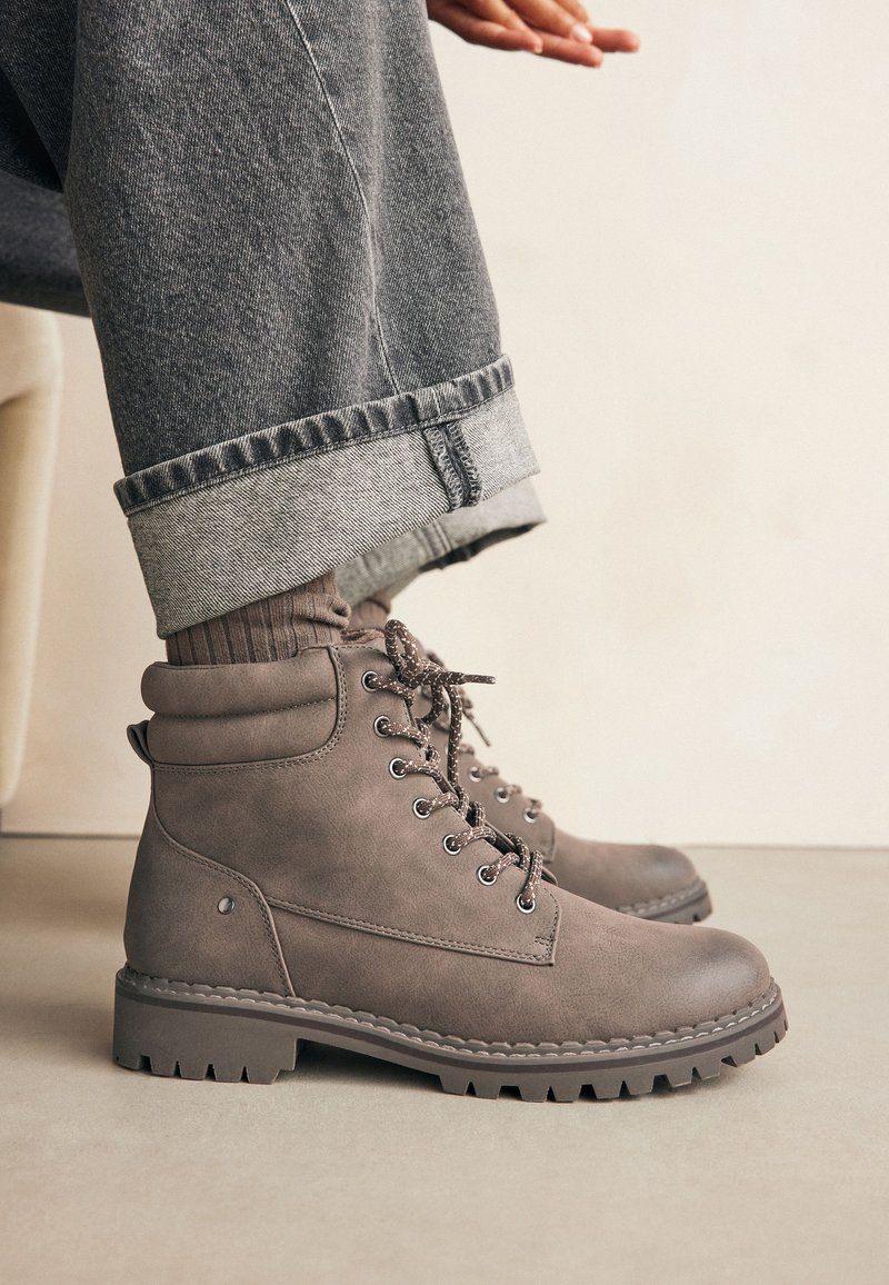 Botas de tobillo marrones hechas de material sintético texturizado, con cordones, ojetes de metal y una suela resistente con un dibujo pronunciado.