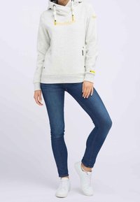 Světle šedá mikina s velkým stojáčkem, šňůrkami a žlutým akcentem. V kombinaci se štíhlými modrými džíny a bílými teniskami.