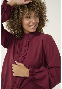 Burgundy blouse made of soft fabric with a ruffled front placket, high collar, and gathered sleeves, featuring button details and a loose fit.