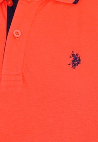 Rød polo skjorte lavet af struktureret stof, med en marineblå krave, to knapper og et lille blåt broderet logo på brystet.