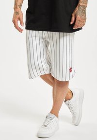 Shorts blancs à rayures pour hommes avec une coupe décontractée, présentant un accent de logo rouge. Associés à des baskets blanches. Le tissu semble léger.