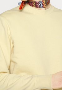 Sudadera amarilla con cuello redondo y mangas largas, que presenta un pequeño logo blanco. Una bufanda naranja y estampada está atada alrededor del cuello.