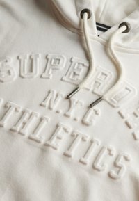 Hvid hættetrøje lavet af blødt stof, med præget tekst "SUPER NYC ATHLETICS" og snore med metaludtræk.