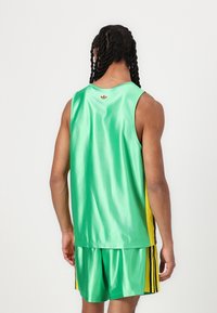 Tricou sportiv fără mâneci, de culoare verde, cu un aspect strălucitor, având accente laterale galbene și dungi negre. Logo mic roșu la gât.
